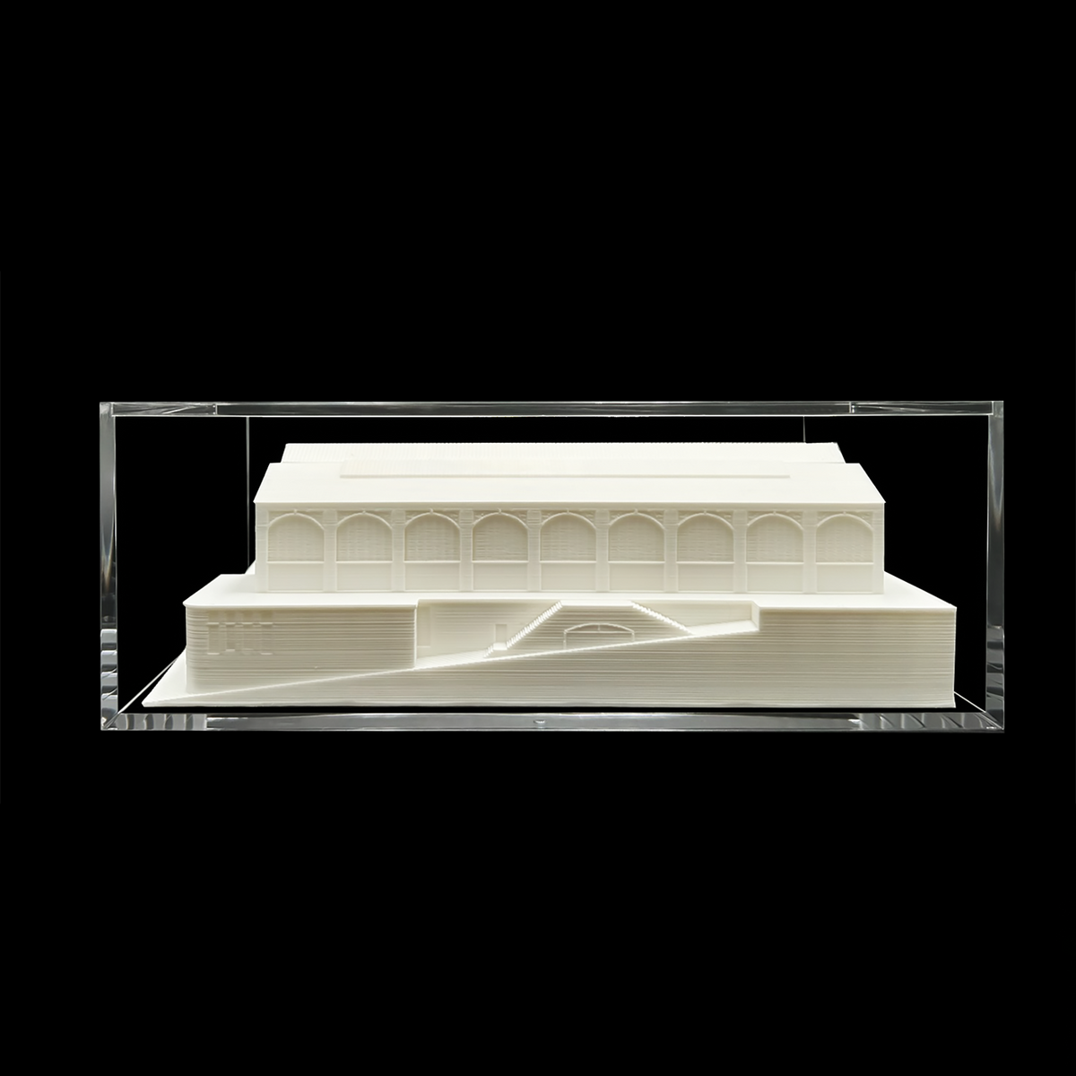 A white architectural model of a building, displayed in a clear glass case against a black background.