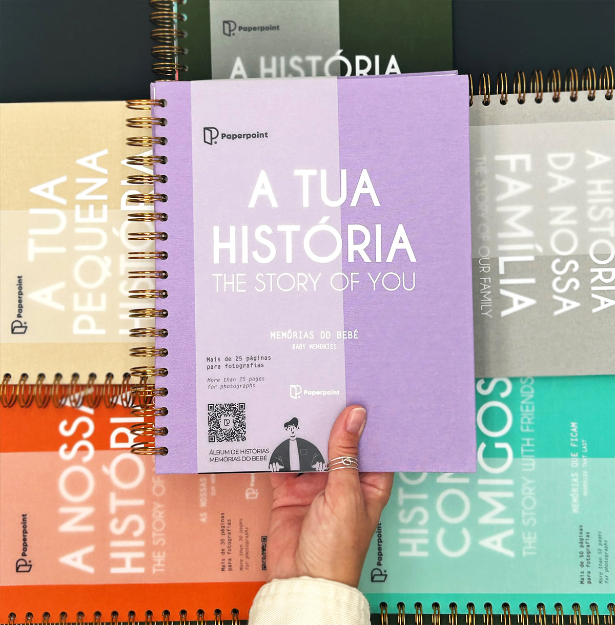 A hand holding a purple book titled "A Tua Historia: The Story of You" in front of a collection of colorful spiral notebooks.
