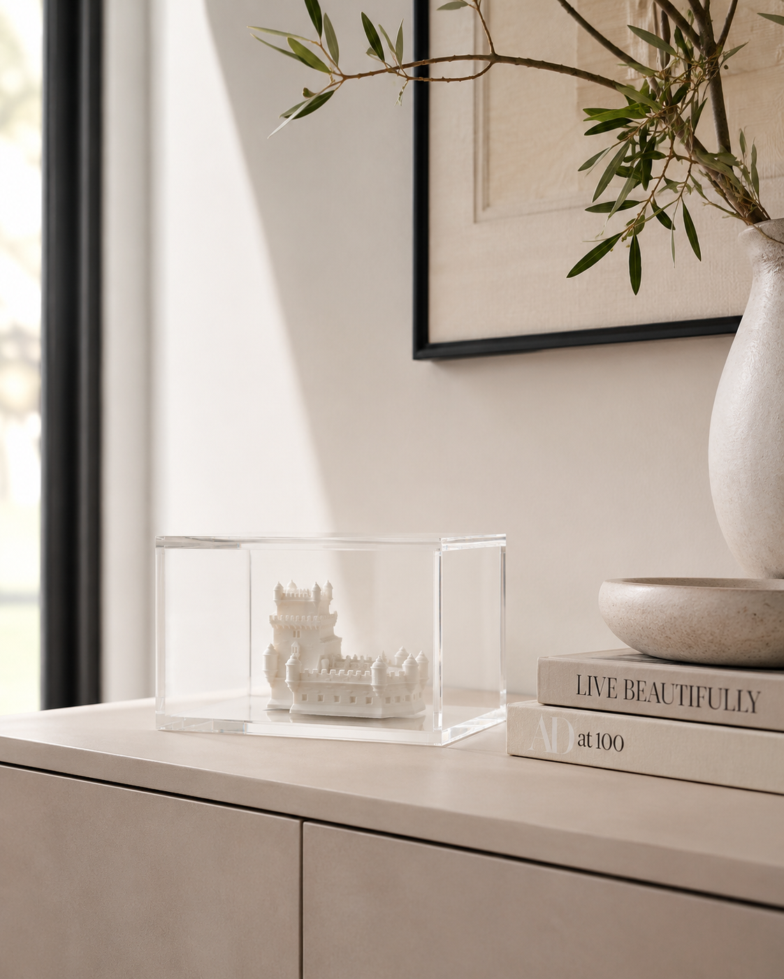 a minimalist interior design with a clear glass box containing a small model ship, a white vase with greenery, and a stack of books titled "Live Beautifully" on a white dresser.