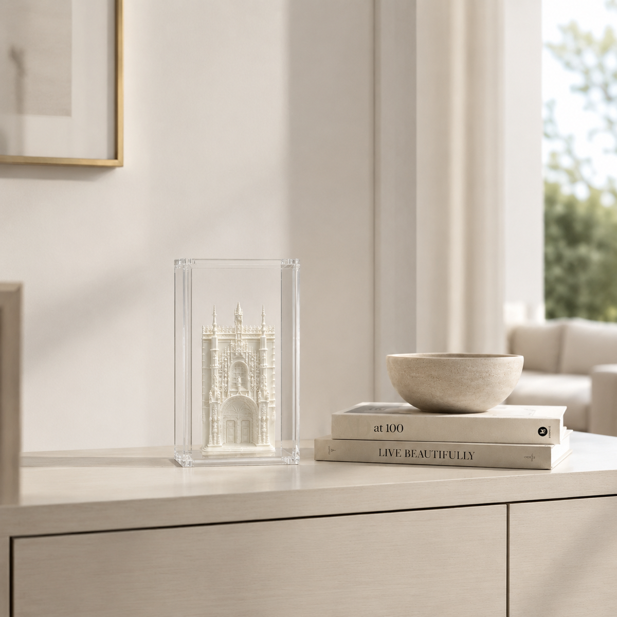 a white cabinet with a clear glass case containing a miniature model of a building, and a stack of books titled "Live Beautifully" on top of the cabinet.