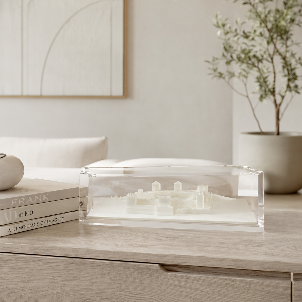 a clear glass display case containing a miniature model of a building, placed on a wooden surface. In the background, there is a potted plant and a stack of books, creating a cozy and inviting atmosphere.