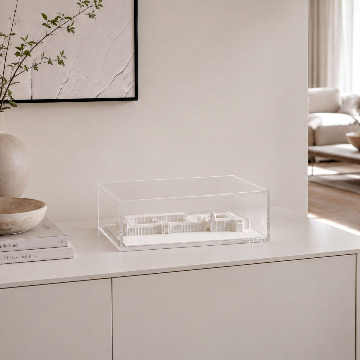 a minimalist interior design with a white dresser, a clear glass box containing a model of a building, and a framed artwork on the wall.