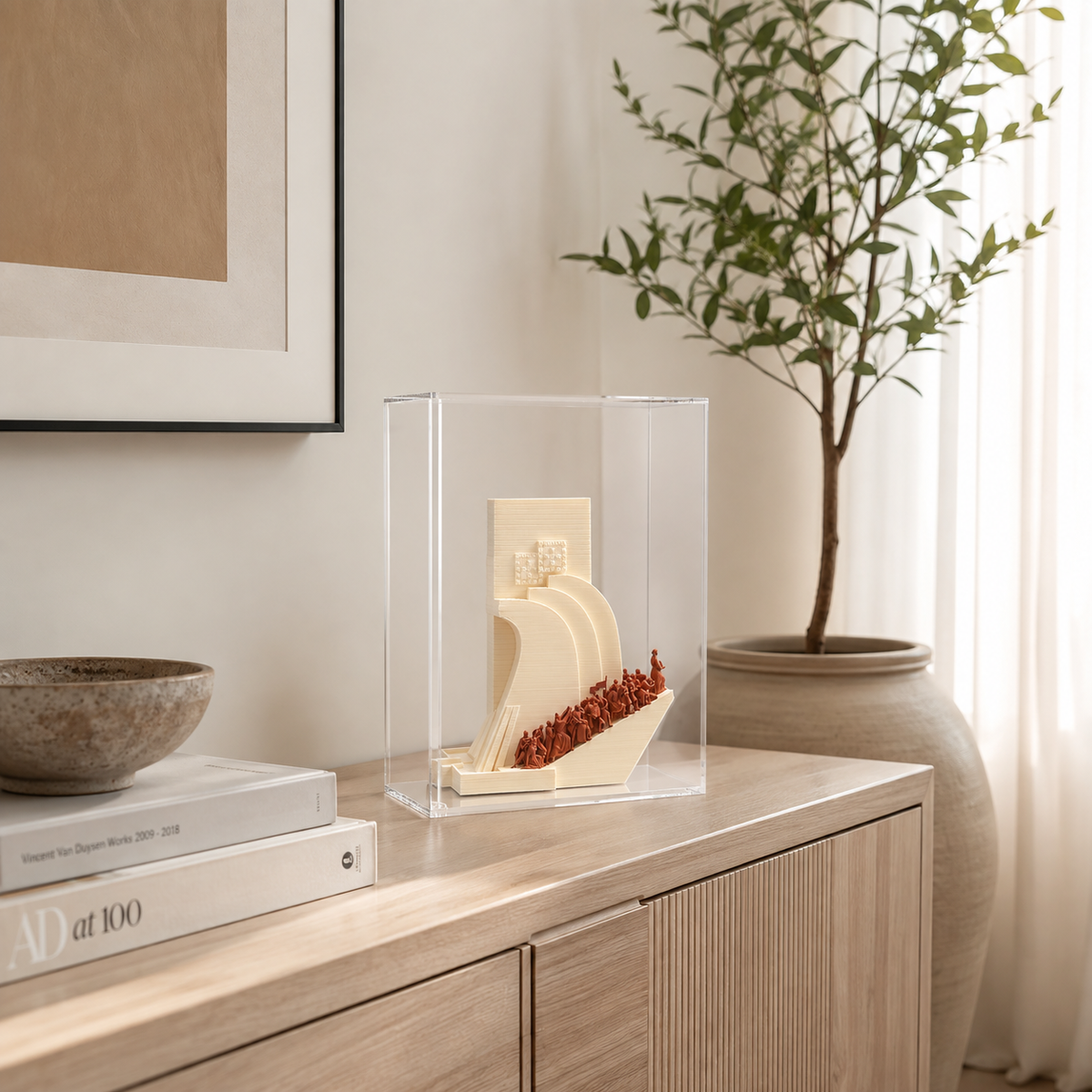 a wooden cabinet with a clear glass display case on top, containing a small sculpture or model. The cabinet is situated next to a large vase with a plant, and there are some books on a nearby surface. The overall scene appears to be a well-organized and aesthetically pleasing display.