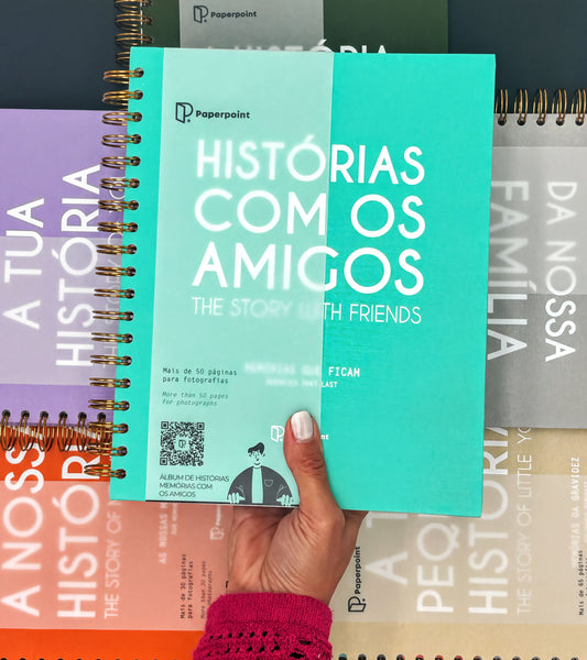 A hand holding a turquoise spiral notebook titled "HISTORIAS COM OS AMIGOS: THE STORY WITH FRIENDS" with a QR code on the cover.