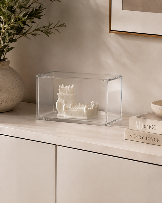 a clear glass display case containing a small white sculpture of a castle, placed on a white cabinet or shelf.