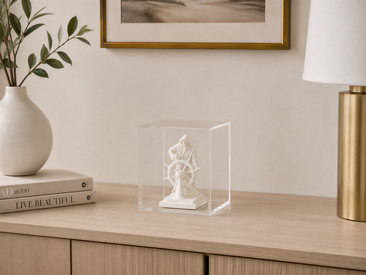 a wooden desk with a clear glass display case containing a small white statue of a person, possibly a sailor, and a stack of books. There is also a lamp and a plant on the desk.