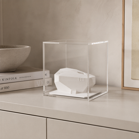 a white ceramic object, possibly a sculpture or a piece of art, displayed in a clear glass case on a white surface.