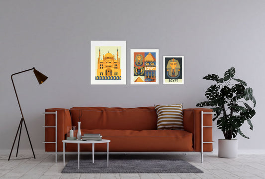 a cozy living room with a brown sofa, a coffee table, and a potted plant. Above the sofa, there are three framed posters depicting Egyptian landmarks and symbols, including a pyramid, a pharaoh, and a temple.