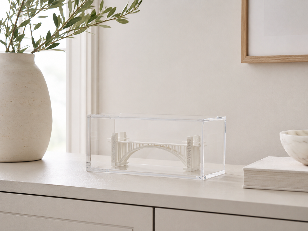 a clear glass box containing a miniature model of a bridge, placed on a white surface next to a vase with green leaves and a framed picture.