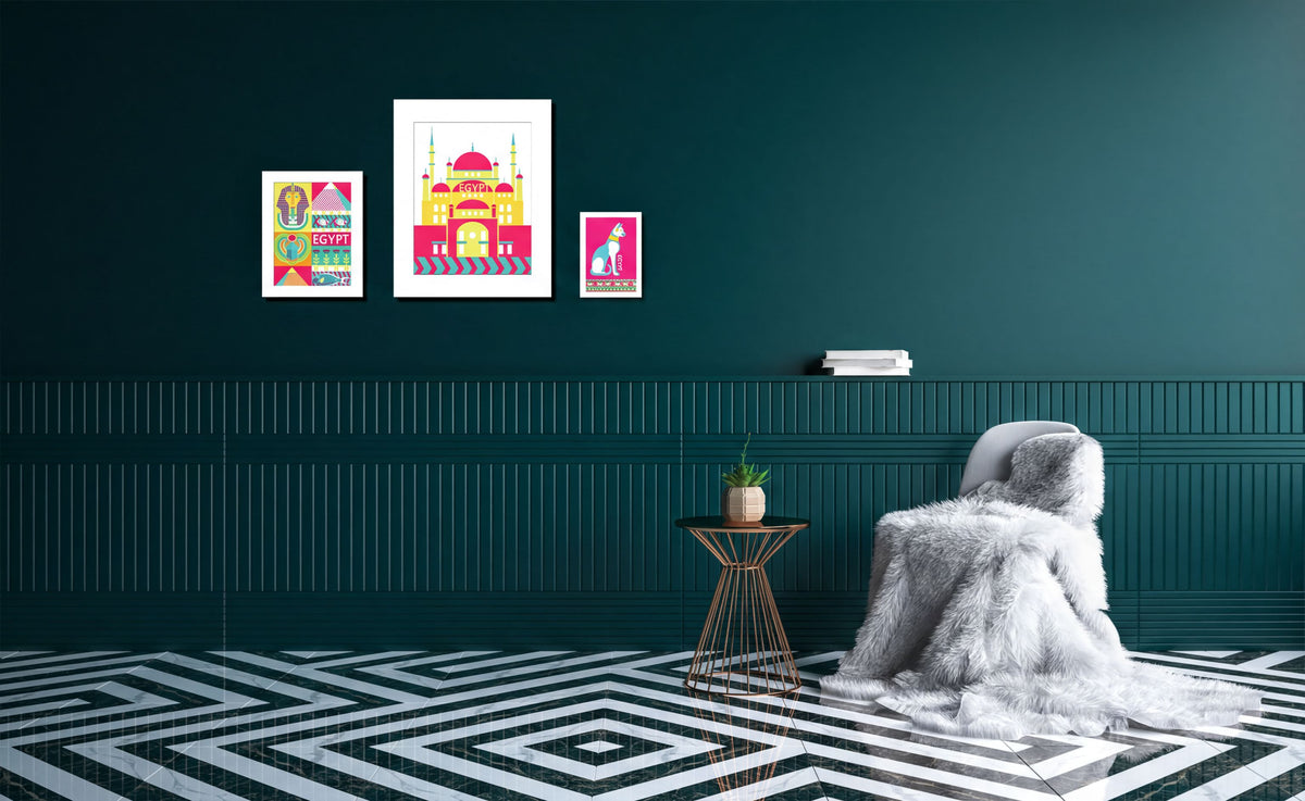 a cozy living room with a plush white blanket draped over a chair, a small side table, and a geometric patterned floor. The walls are painted a dark green color, and there are three framed artworks displayed on the wall, including a colorful illustration of a building and a pink and white illustration of a cat.