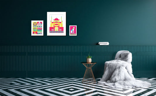 a cozy living room with a plush white blanket draped over a chair, a small side table, and a geometric patterned floor. The walls are painted a dark green color, and there are three framed artworks displayed on the wall, including a colorful illustration of a building and a pink and white illustration of a cat.