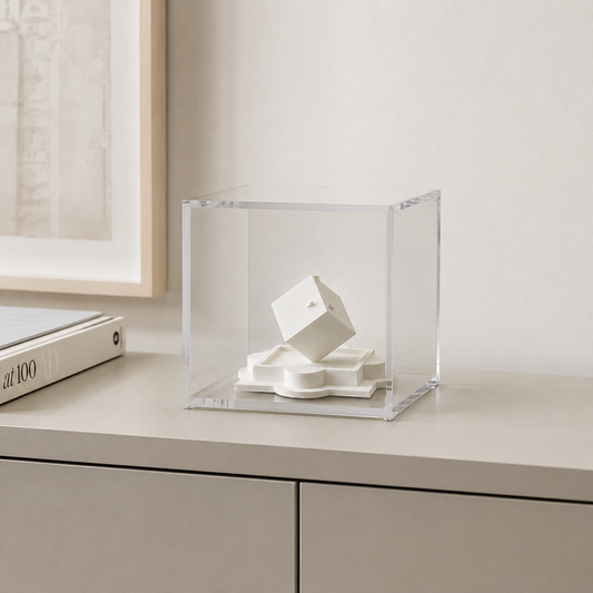 A white cube sculpture is displayed in a clear glass case on a white surface, with a framed picture visible in the background.