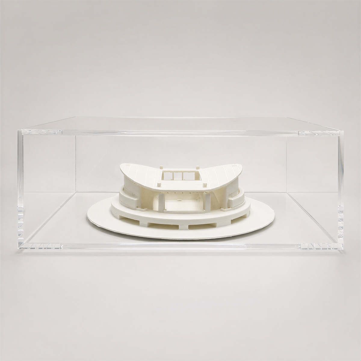 A white, circular model of a building or structure is displayed in a clear, rectangular glass case.