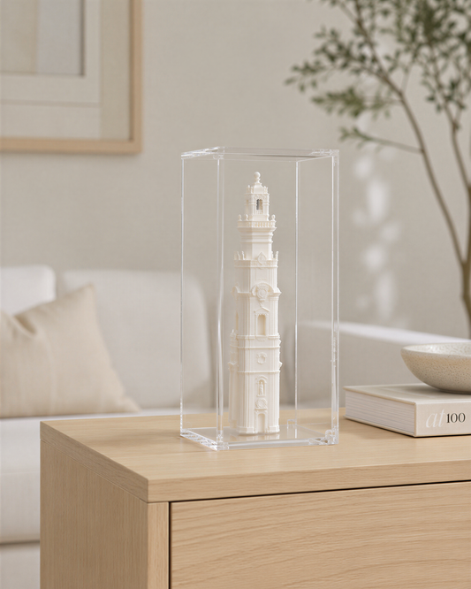 A small white tower model is displayed in a clear glass case on a wooden table, with a plant and a book visible in the background.
