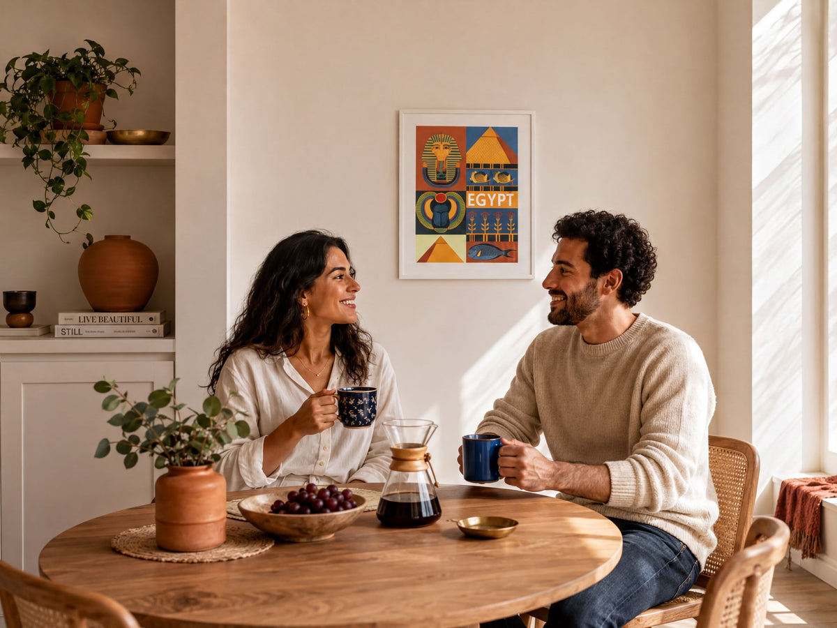 A man and a woman are sitting at a wooden table, engaged in conversation while holding coffee cups. The table is set with a vase of flowers, a bowl of fruit, and a pitcher of water. A framed poster of Egypt is displayed on the wall behind them.
