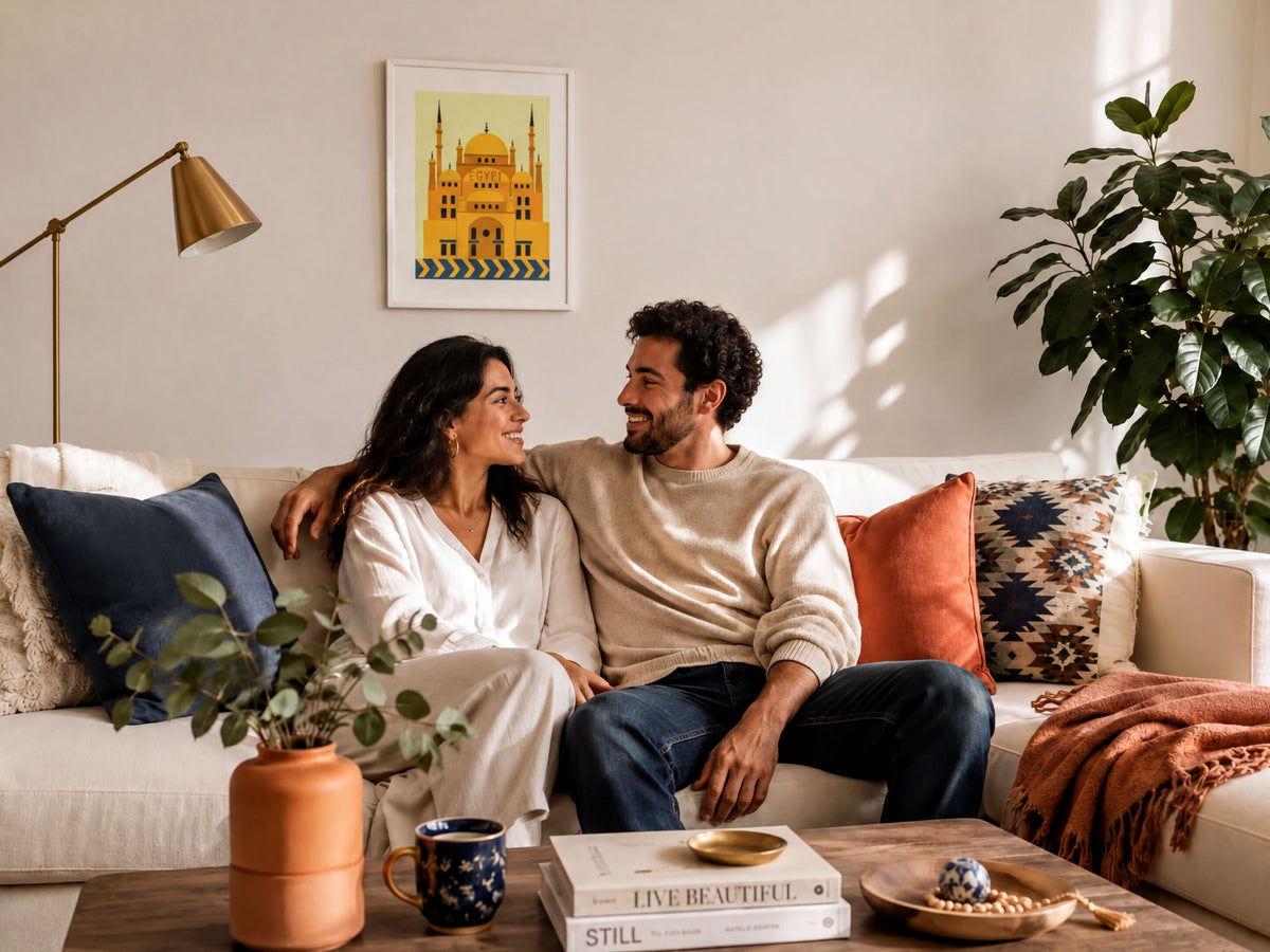A couple sits together on a couch, smiling and looking at each other, with a framed picture of a building on the wall behind them.