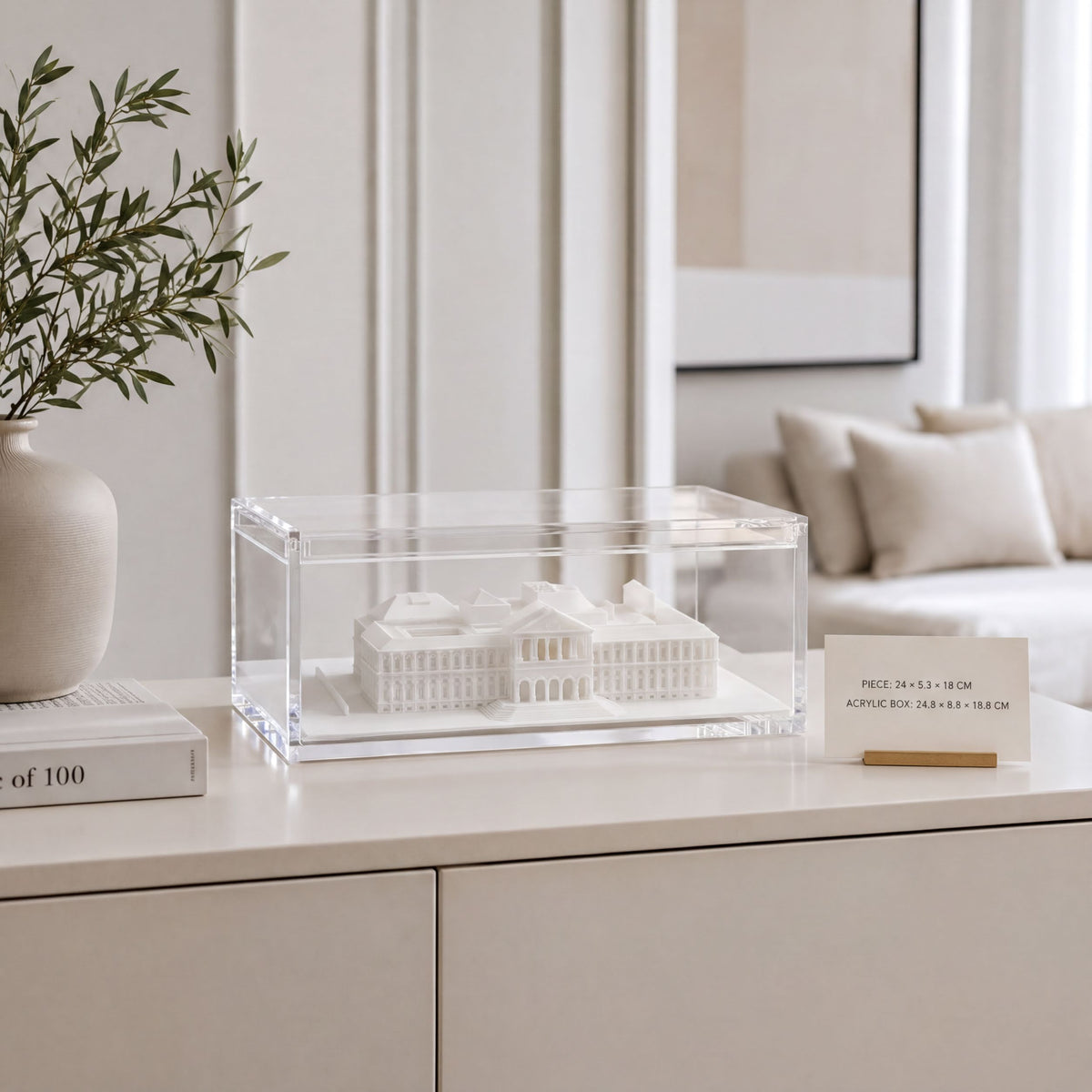 a white model of a building displayed in a clear case on a white surface, with a vase of greenery and a card with text in the background.