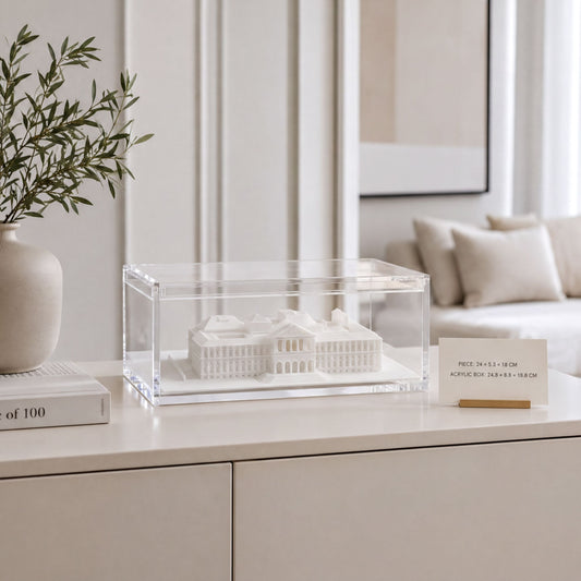 a white model of a building displayed in a clear case on a white surface, with a vase of greenery and a card with text in the background.
