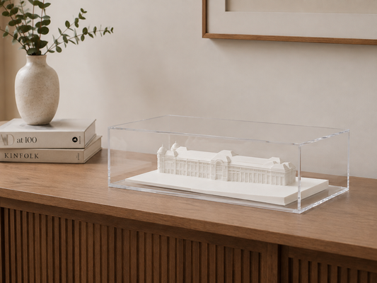 a wooden table with a clear glass case containing a white architectural model of a building, placed next to a stack of books and a vase with greenery.