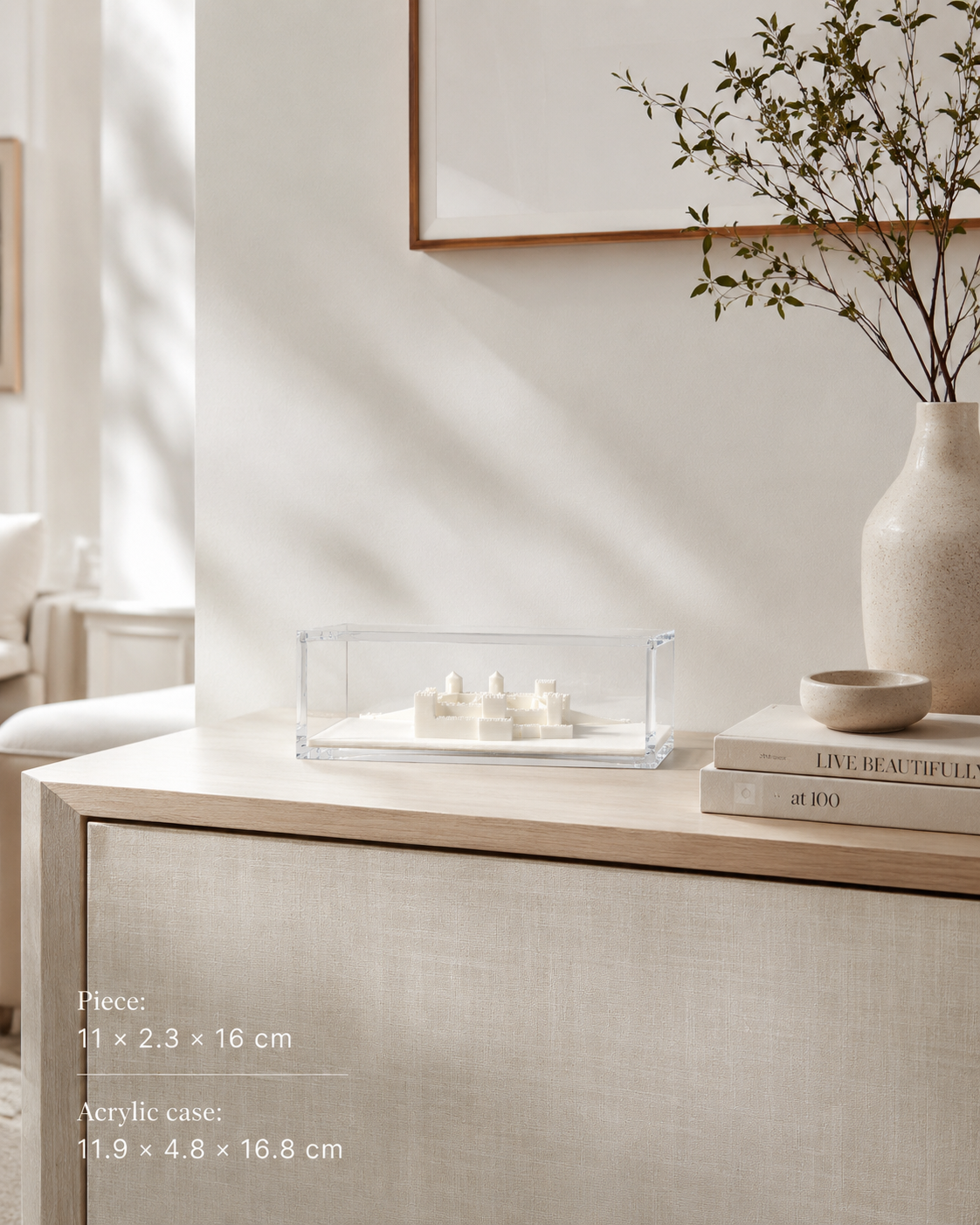 a minimalist and modern interior design with a clear acrylic case containing small white objects, a white vase with greenery, and a stack of books on a wooden table.