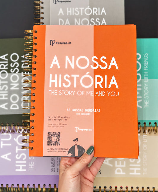 A hand holding an orange spiral notebook with the title "A NOSSÁ HISTORIA" printed on it.