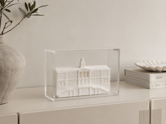 a clear glass display case containing a miniature model of a building, placed on a white surface next to a vase and some books.