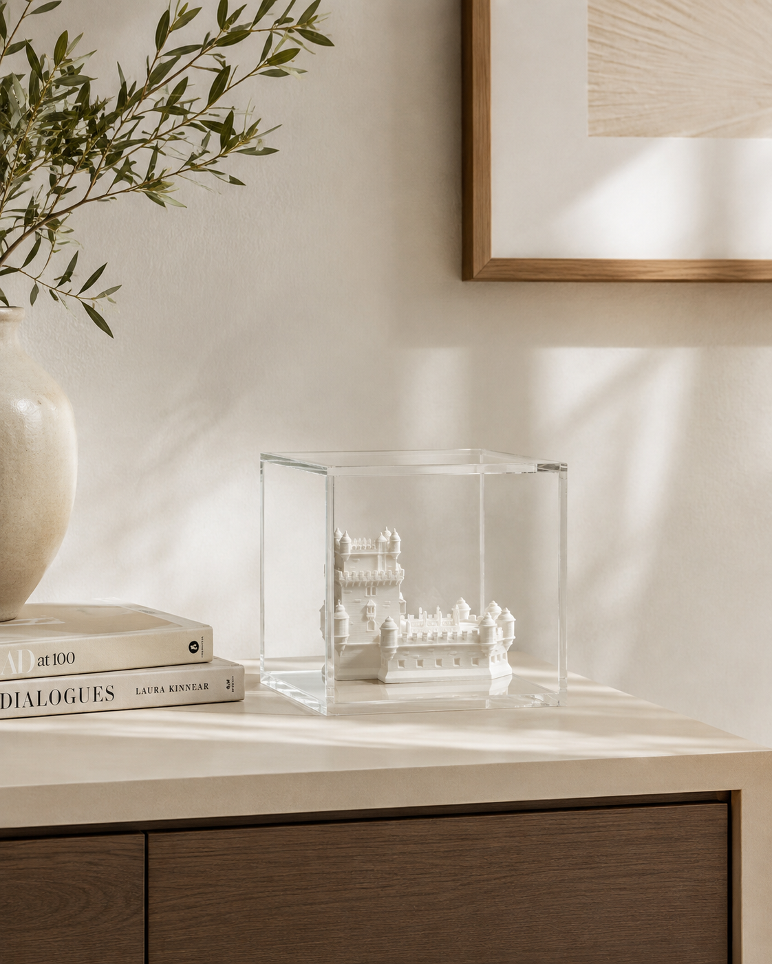 a glass display case containing a small white sculpture, placed on a wooden surface next to a stack of books and a white vase.