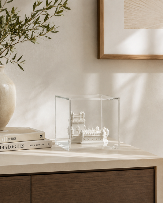 a glass display case containing a small white sculpture, placed on a wooden surface next to a stack of books and a white vase.