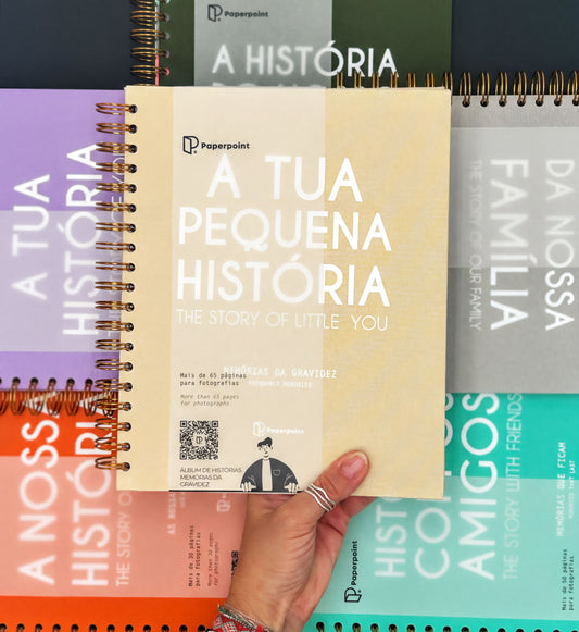 A hand holding a book titled "A Tua Pequena Historia: The Story of Little You" in front of a collection of other books.
