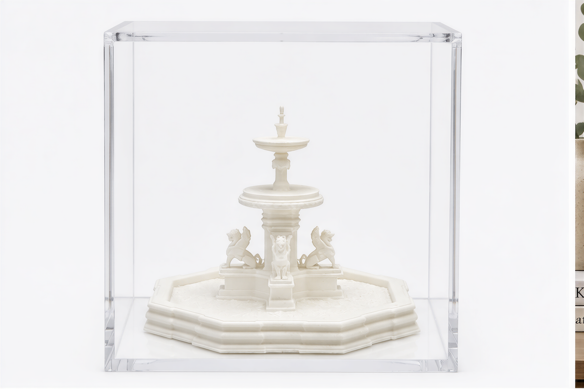A white ceramic fountain with a central spout and two lion-like figures on either side is displayed in a clear glass case.