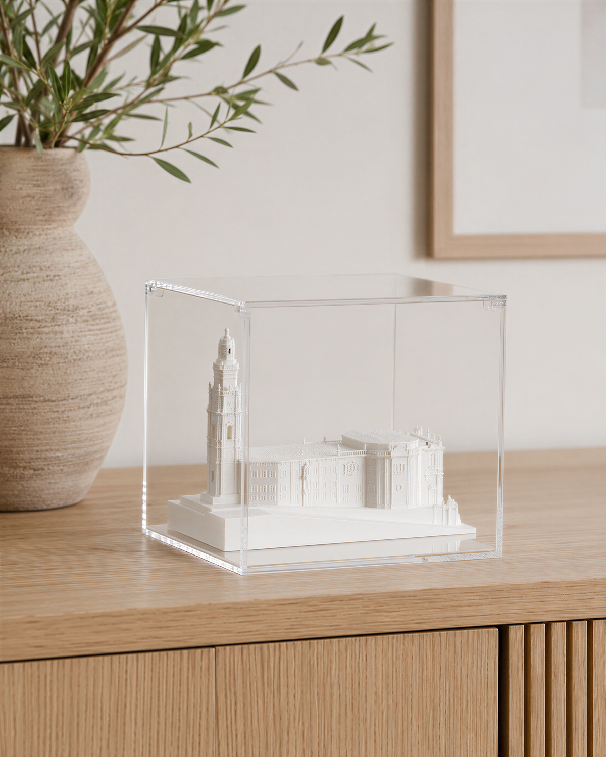 A small white model of a building is displayed in a clear glass case on a wooden surface, with a vase of greenery to the left.