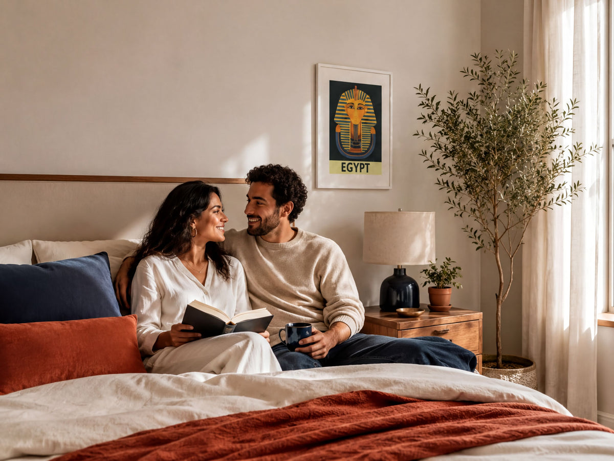 A couple sits together on a bed, with the woman reading a book and the man looking at her with a smile.