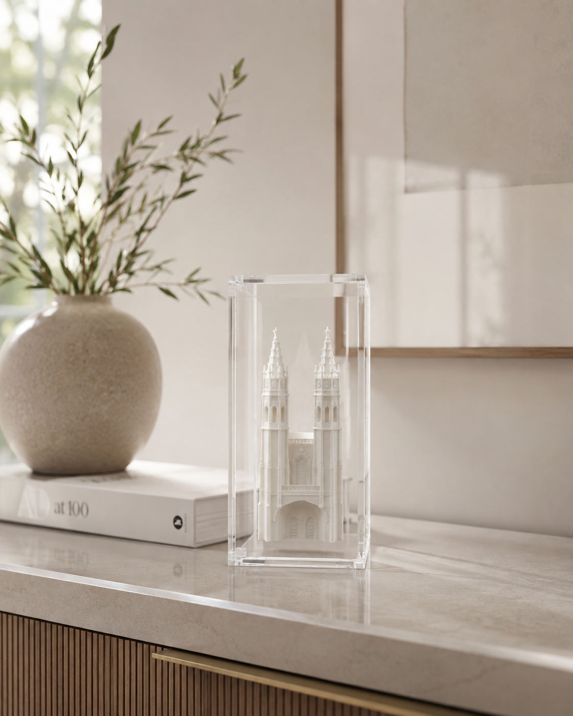 a clear glass box containing a miniature model of a castle, placed on a marble countertop next to a book and a vase with greenery.
